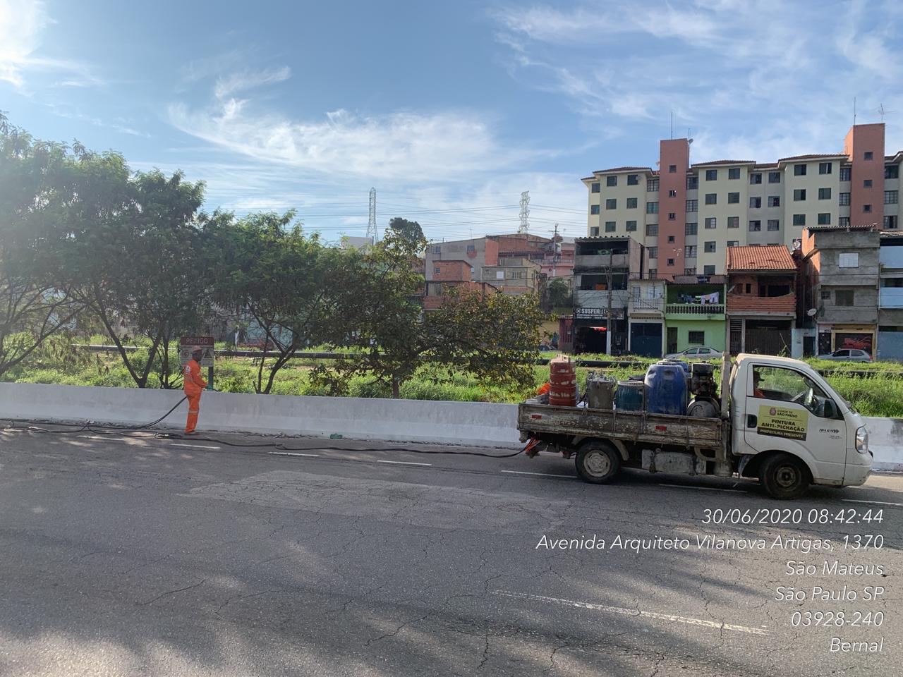 Caminhão de pintura a jato está parado diante de avenida, com prédios de apartamentos ao fundo. Um funcionário de macacão laranja vai em direção ao caminhão. 